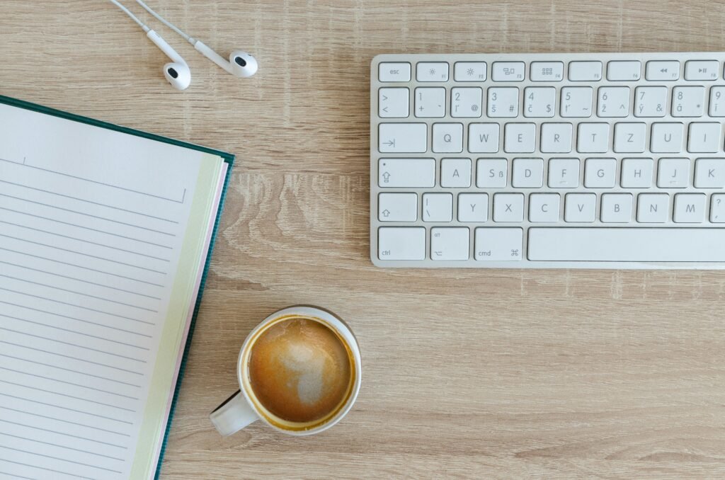 Top-down view of a desk with a notebook, white keyboard, white earbuds, and a cup of coffee