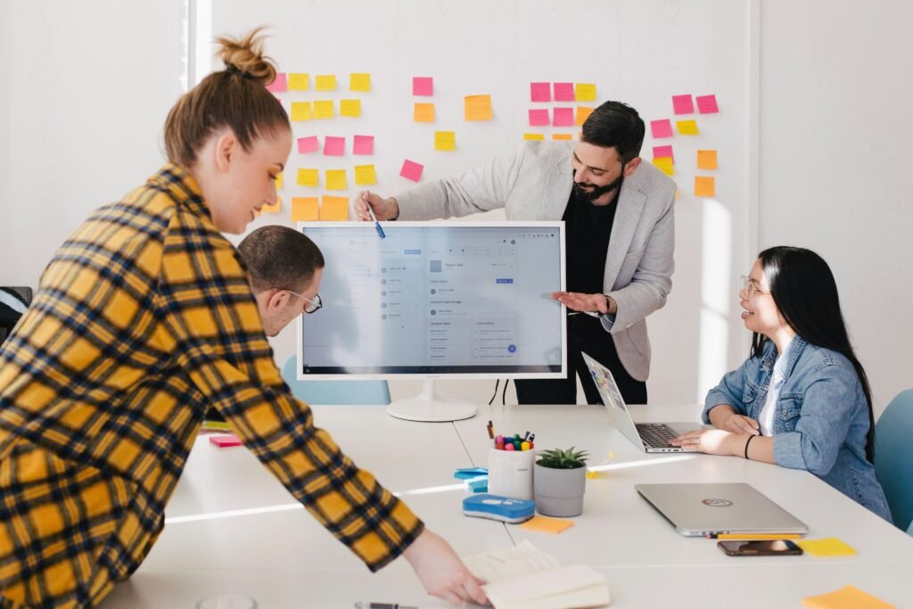 Four colleagues in a bright office discussing a project around a large monitor, with colorful sticky notes on the walls.