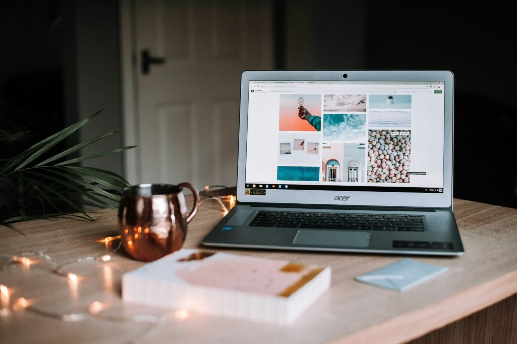 A cozy, softly lit workspace featuring an open Acer laptop displaying a Pinterest-style mood board of pastel-toned photos. The wooden desk also holds a copper mug, a palm frond plant, a blurred book in the foreground, fairy lights, and a light blue envelope, creating a warm and aesthetically pleasing environment ideal for creative work or inspiration browsing.