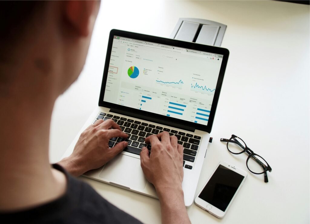 A person types on a laptop showing a Google Analytics dashboard with graphs, charts, and metrics. A smartphone and a pair of glasses are placed nearby on the white desk, indicating data-driven work in a digital marketing or analytics setting.