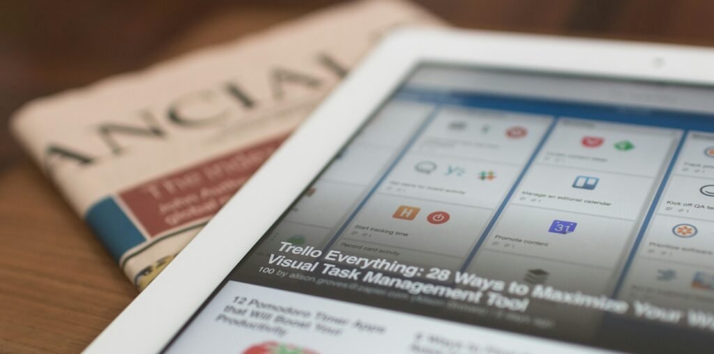 A white tablet displaying a Trello dashboard article titled "Trello Everything: 28 Ways to Maximize Your Visual Task Management Tool" rests on top of a folded newspaper with a partially visible headline reading "ANCIAL." The image suggests productivity and staying informed through both digital and print media.